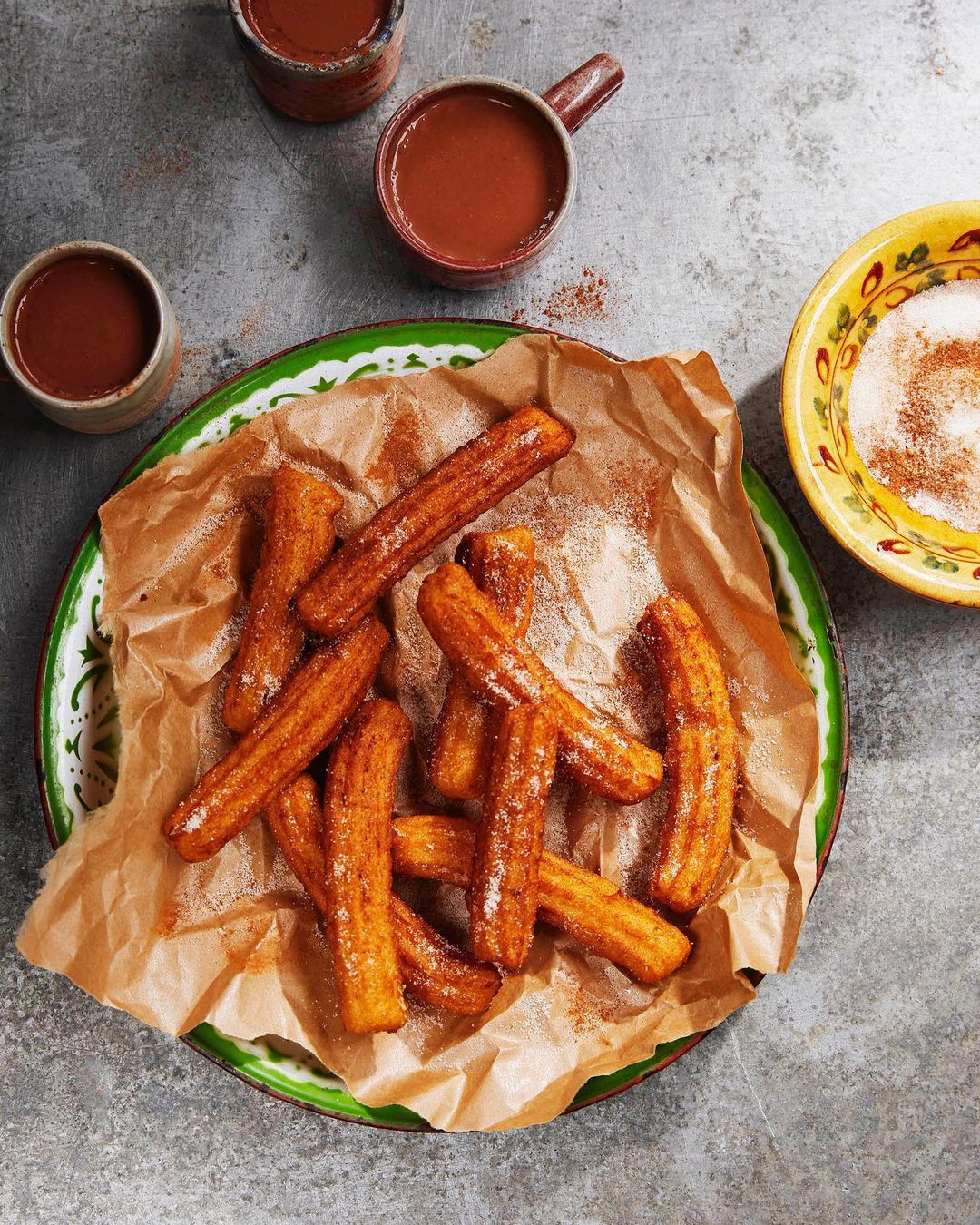 Overhead shot features a plate of churros dusted with sugar, accompanied by mugs of hot chocolate and a bowl of sugar for dipping.