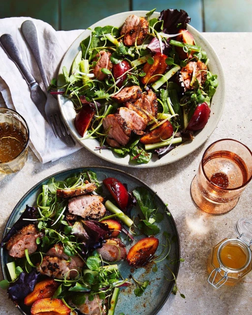 Overhead shot of two plates of salad with greens, meat, and sliced plums; one with silverware and beverages, food photography.