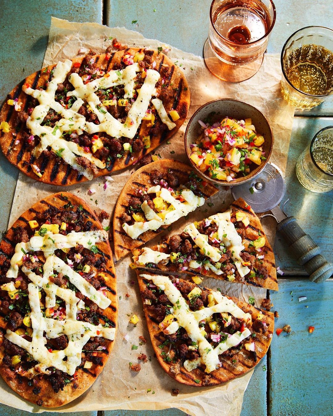 Flatbread pizzas with grilled marks, topped with cheese and savory ingredients next to a bowl of salsa and a couple of drinks.