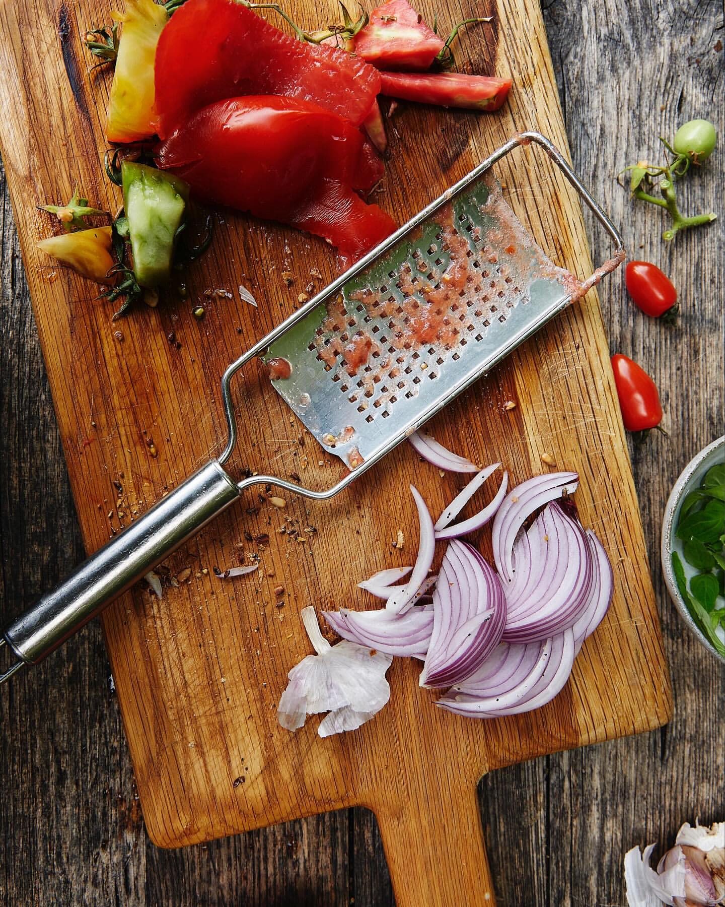 Overhead view of a wooden cutting board with red tomatoes, a metal grater, and sliced red onions, shot with natural lighting.