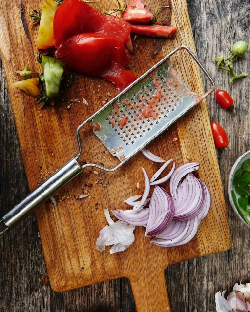 Overhead view of a wooden cutting board with red tomatoes, a metal grater, and sliced red onions, shot with natural lighting.
