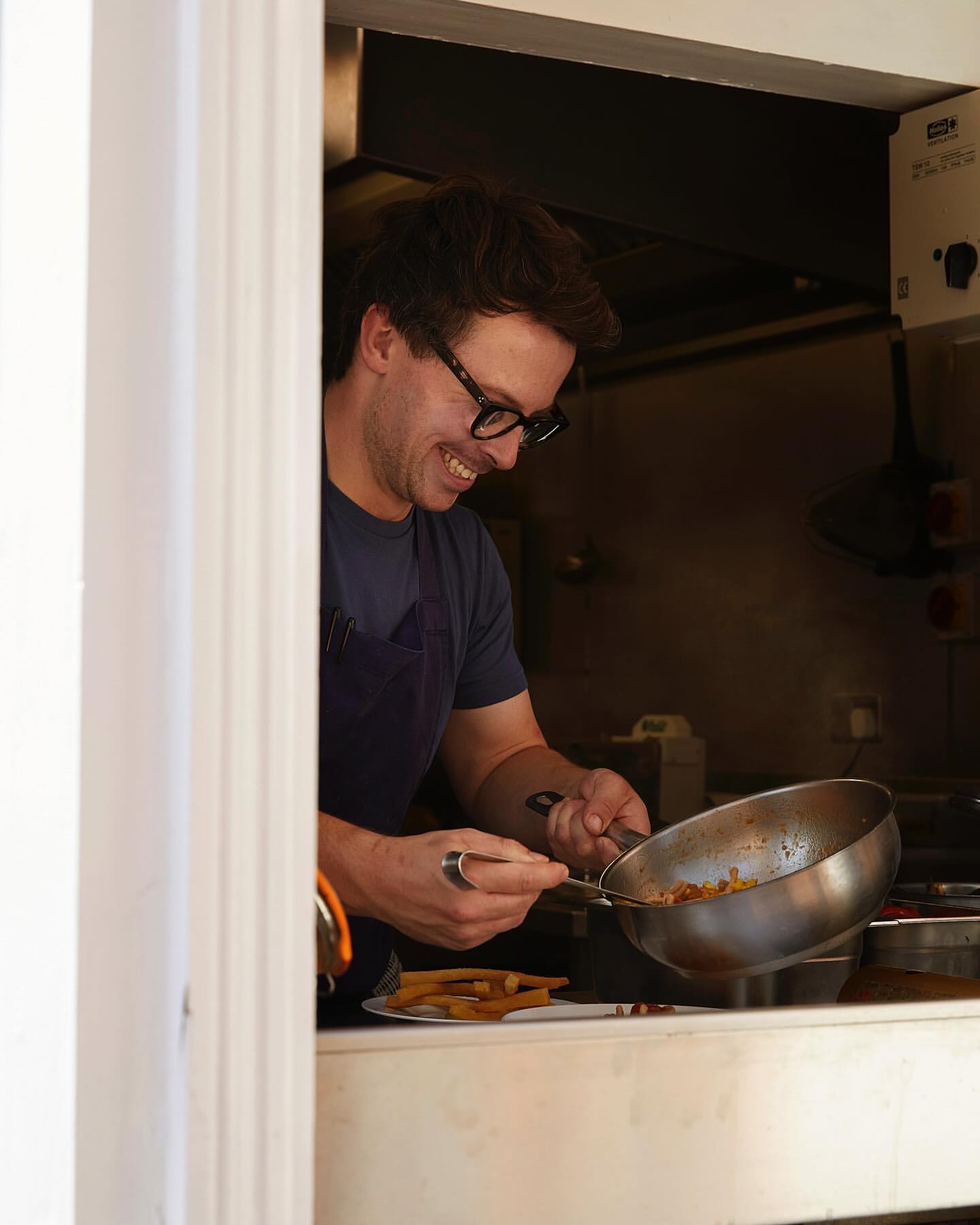 Smiling chef prepares food in a metal pan in a restaurant kitchen, visible through a serving window, showcasing culinary skill and cuisine.