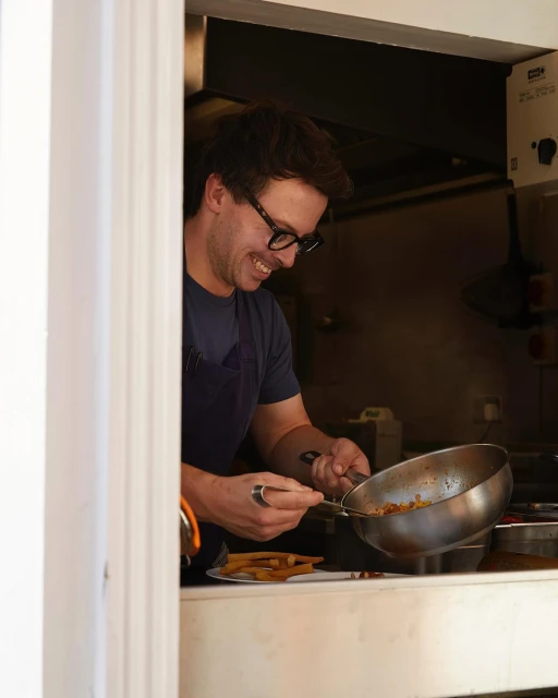 Smiling chef prepares food in a metal pan in a restaurant kitchen, visible through a serving window, showcasing culinary skill and cuisine.