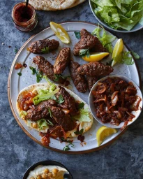 Top down overhead food photography of spiced minced lamb kofte served with flatbread, lemon wedges, and caramelized onions on a dark slate surface.