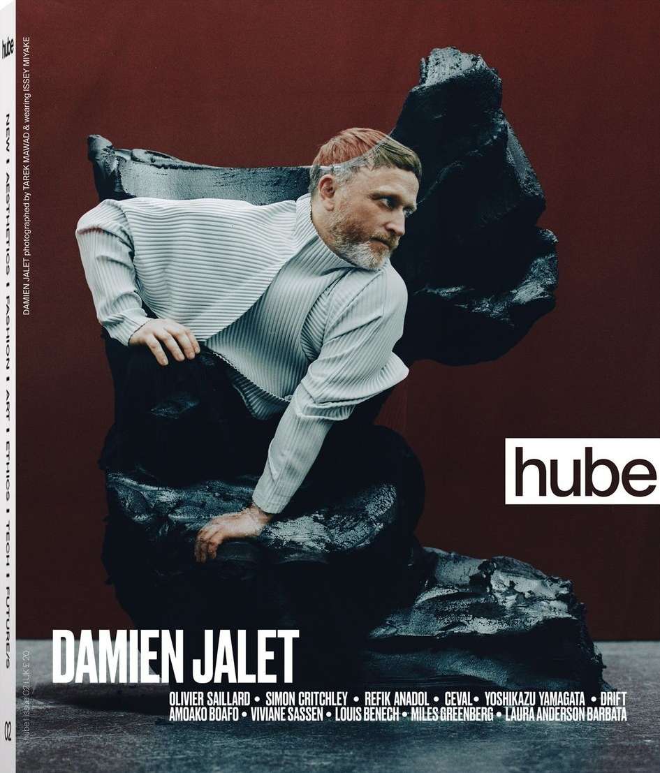 Fashion magazine cover featuring an adult male model in a striped shirt and dark pants sitting on a textured black sculpture with a red backdrop.