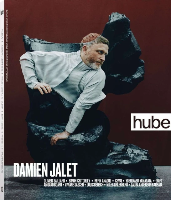 Fashion magazine cover featuring an adult male model in a striped shirt and dark pants sitting on a textured black sculpture with a red backdrop.