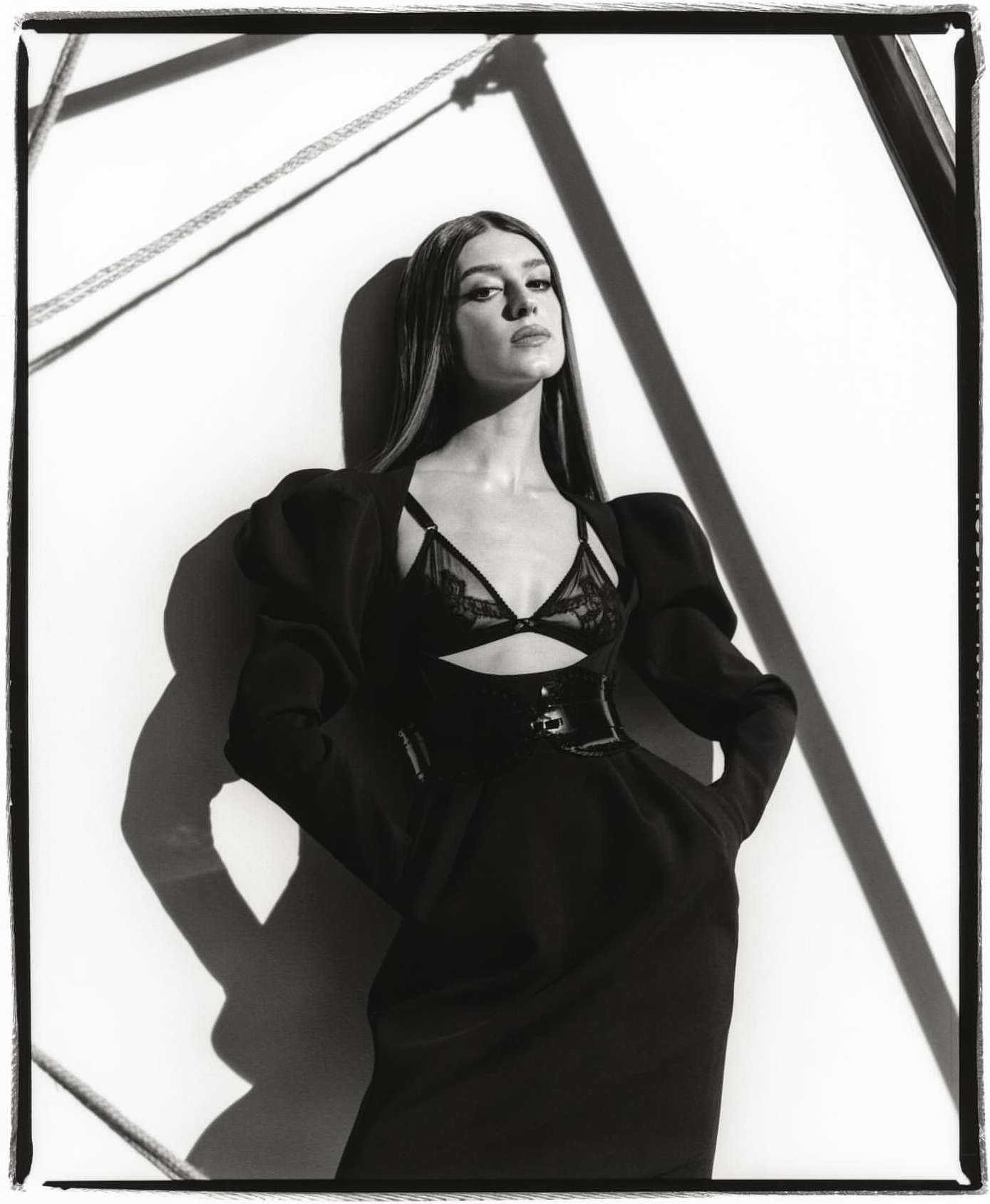 Black-and-white studio shot of a woman in a black dress with a lace bralette, dramatic shadows, and geometric background art.