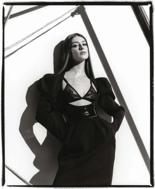 Black-and-white studio shot of a woman in a black dress with a lace bralette, dramatic shadows, and geometric background art.