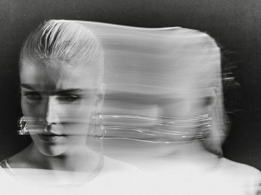 Monochrome portrait of a blonde woman with motion blur and light trails around her face, wearing earrings, against a dark background.