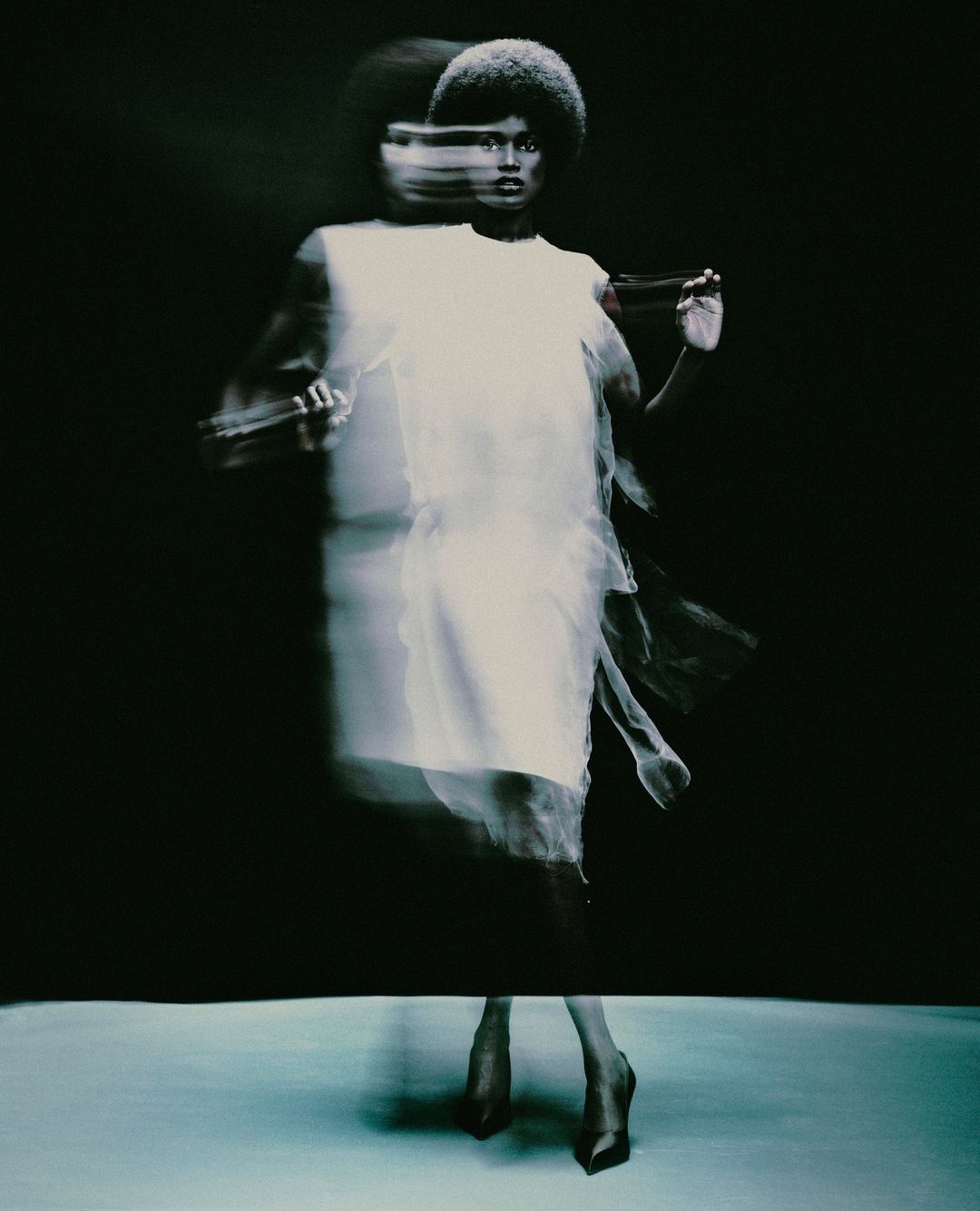 A woman with an afro hairstyle poses in a white dress with a motion blur effect on a neutral gray surface against a dark background.