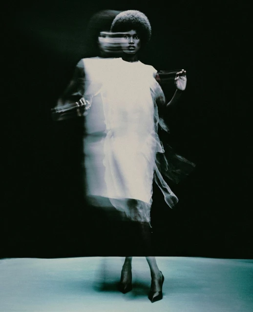 A woman with an afro hairstyle poses in a white dress with a motion blur effect on a neutral gray surface against a dark background.
