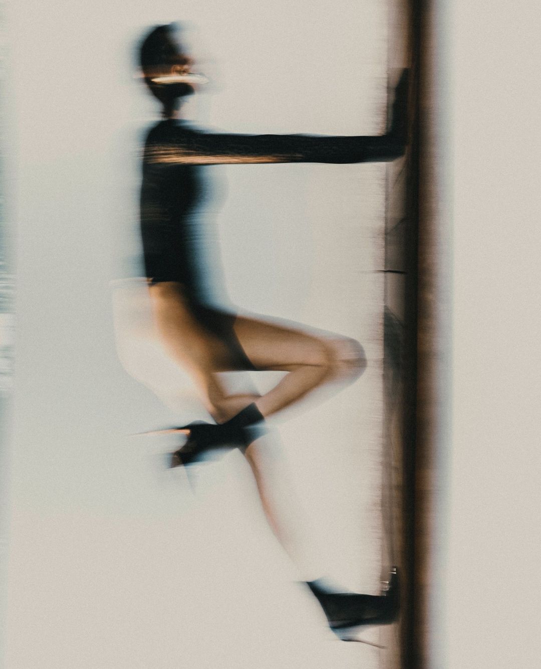 Motion-blurred fashion shot of a woman in black top, shorts, and high heels, leaning against a dark vertical bar with a light background.