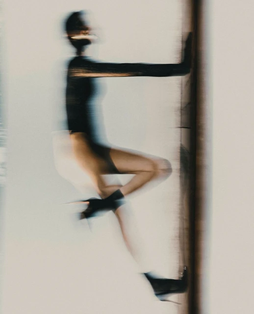 Motion-blurred fashion shot of a woman in black top, shorts, and high heels, leaning against a dark vertical bar with a light background.