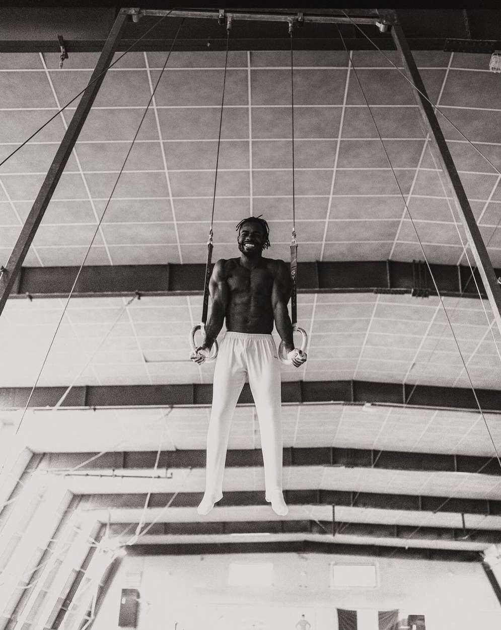 A muscular man with a beard on gymnastic rings, wearing white pants, in a monochrome shot inside a bright indoor athletic facility.