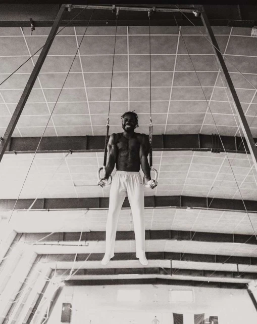 A muscular man with a beard on gymnastic rings, wearing white pants, in a monochrome shot inside a bright indoor athletic facility.
