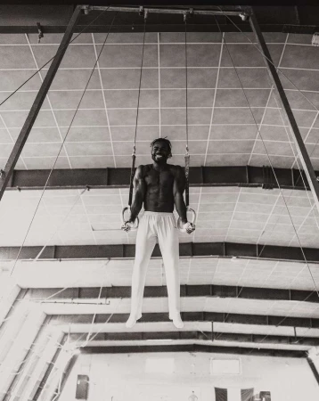 A muscular man with a beard on gymnastic rings, wearing white pants, in a monochrome shot inside a bright indoor athletic facility.