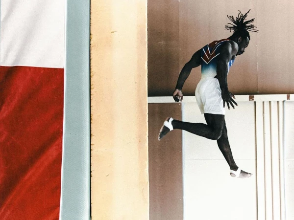 A dark-skinned male gymnast in white shorts and a patterned top is captured mid-air during a floor routine in an indoor gymnasium.