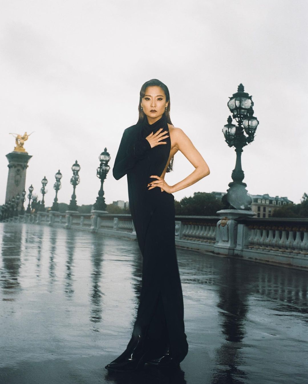 An Asian woman stands on a bridge in a long black dress and one long sleeve, posing with her hand on her chest.