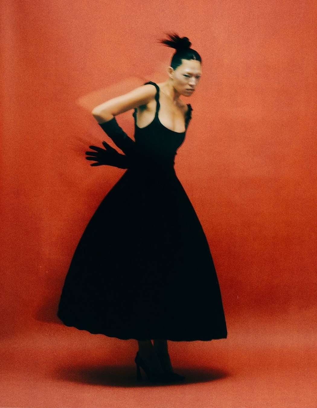 Elegant woman in black dress and gloves poses against a red studio backdrop in a fashion shot featuring motion blur.