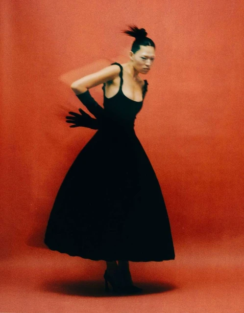 Elegant woman in black dress and gloves poses against a red studio backdrop in a fashion shot featuring motion blur.