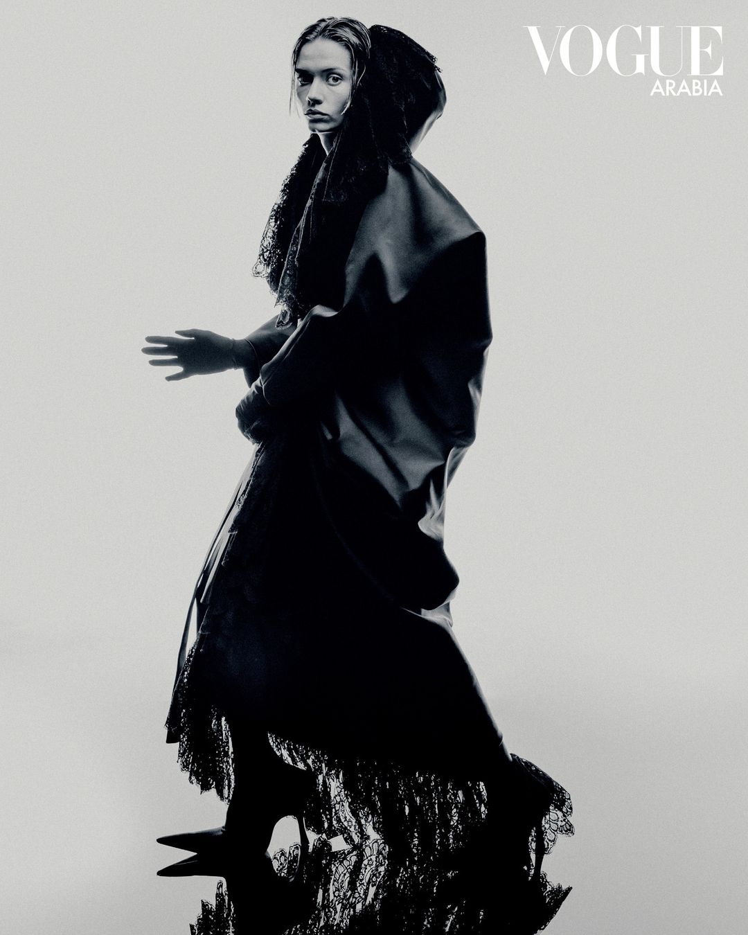 Monochrome fashion shot of a woman in a dark hooded dress standing in a studio setting with Vogue Arabia logo visible in the upper right.