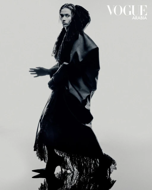 Monochrome fashion shot of a woman in a dark hooded dress standing in a studio setting with Vogue Arabia logo visible in the upper right.