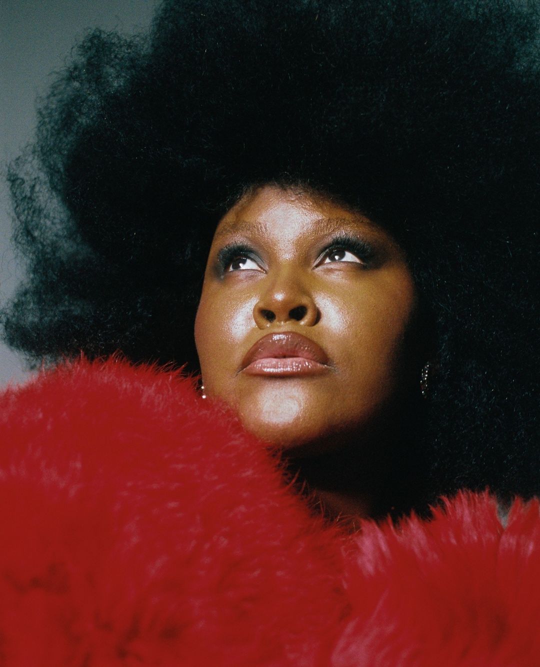 Portrait of a black woman with a large afro and red furry shawl looking up in a photo studio with a gray background.