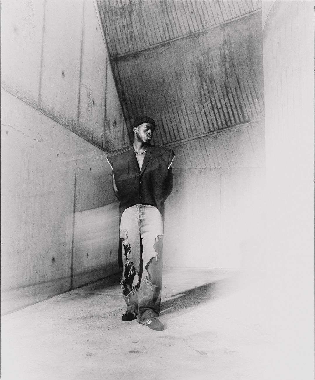 Monochrome portrait of a Black male in a dark vest and ripped jeans, standing in a raw concrete corner, gazing towards the light.