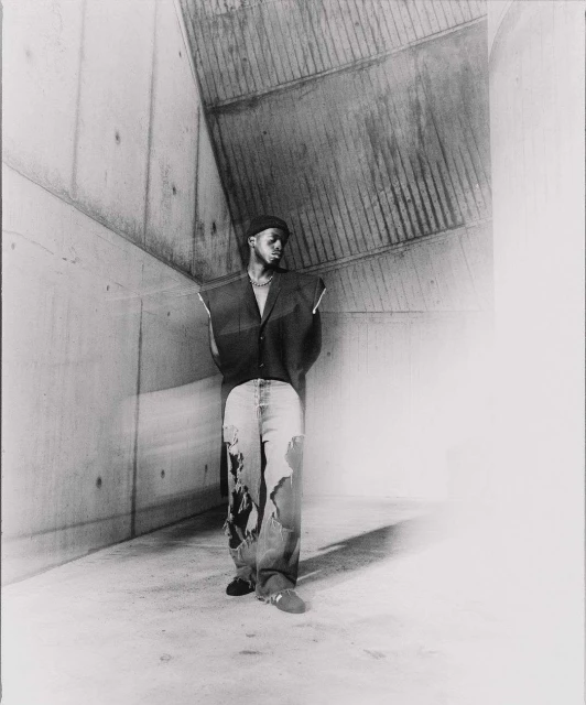 Monochrome portrait of a Black male in a dark vest and ripped jeans, standing in a raw concrete corner, gazing towards the light.