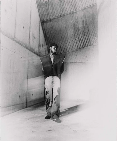 Monochrome portrait of a Black male in a dark vest and ripped jeans, standing in a raw concrete corner, gazing towards the light.
