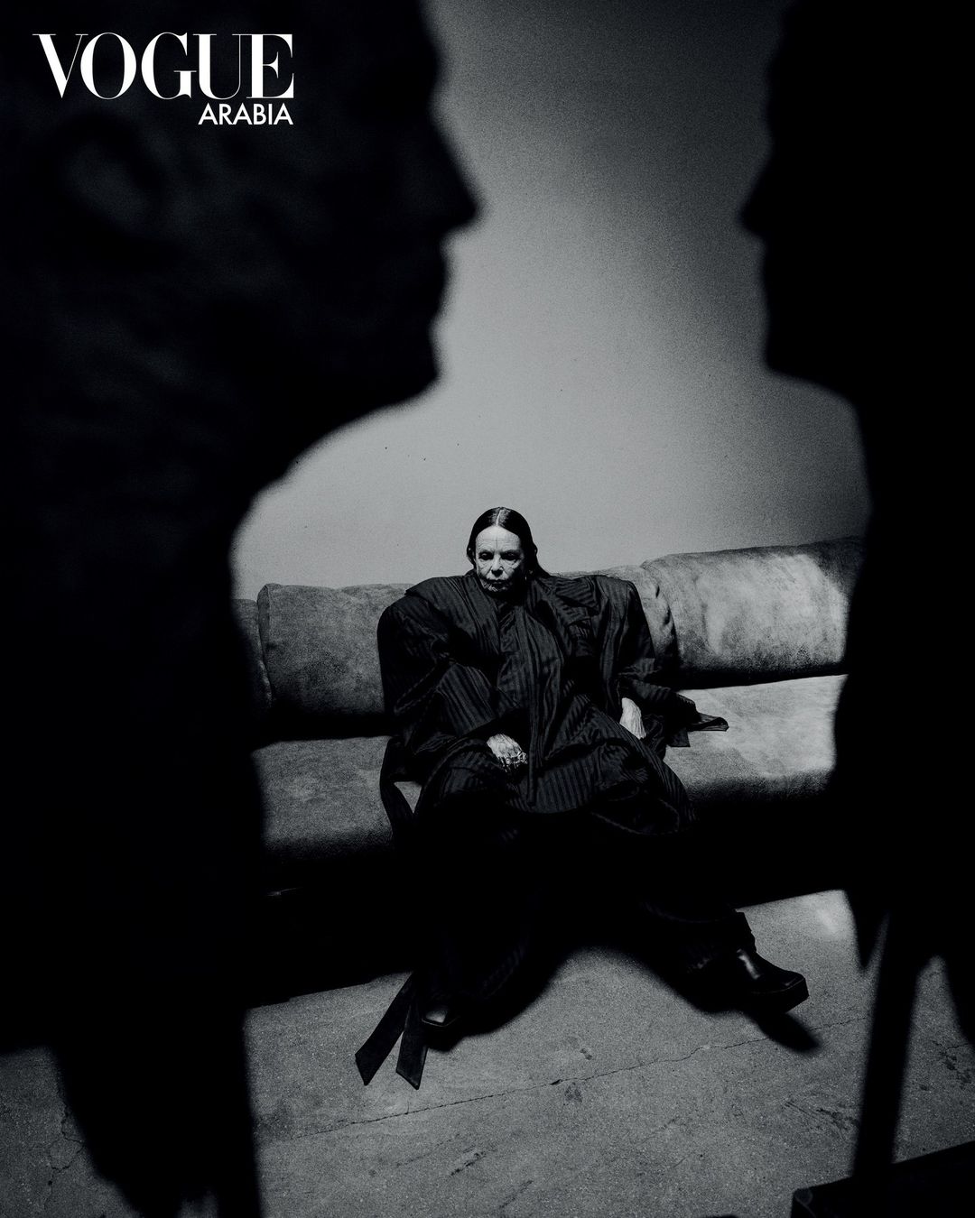 Fashion shot of a woman sitting on a sofa, framed by two shadowy figures, with 'Vogue Arabia' logo visible; black and white photography.