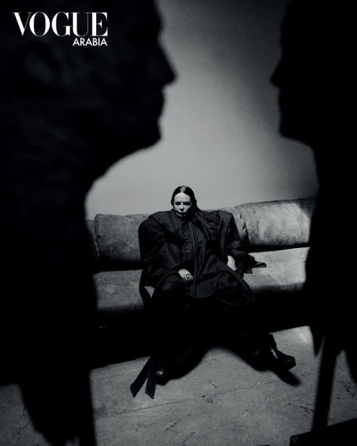 Fashion shot of a woman sitting on a sofa, framed by two shadowy figures, with 'Vogue Arabia' logo visible; black and white photography.