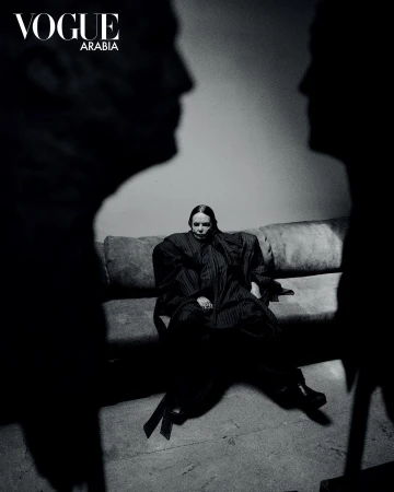 Fashion shot of a woman sitting on a sofa, framed by two shadowy figures, with 'Vogue Arabia' logo visible; black and white photography.