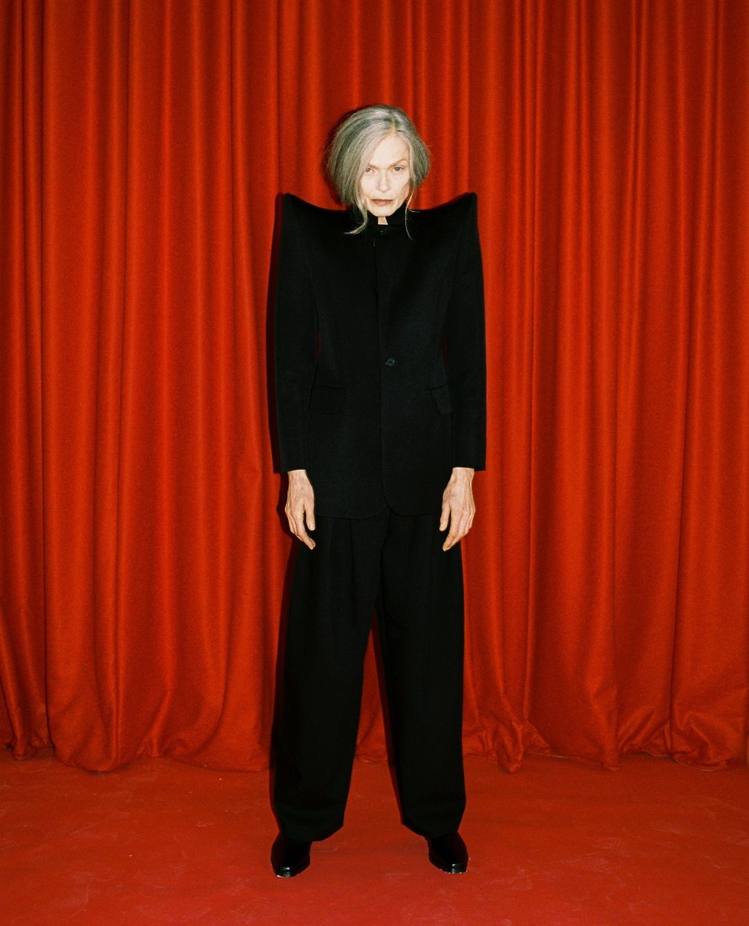 Elegant woman with gray hair, wearing an oversized black blazer and wide-leg pants, poses in front of a vibrant red curtain backdrop.