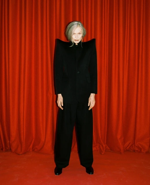 Elegant woman with gray hair, wearing an oversized black blazer and wide-leg pants, poses in front of a vibrant red curtain backdrop.