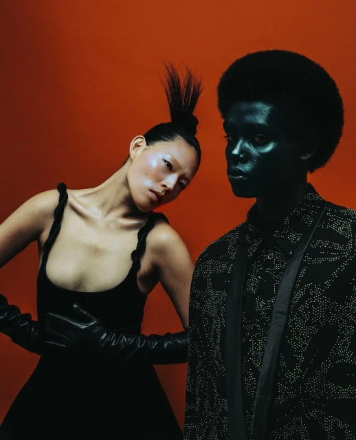 Two fashion models pose in front of an orange backdrop. The female model wears a black dress and gloves, while the male model is painted dark.