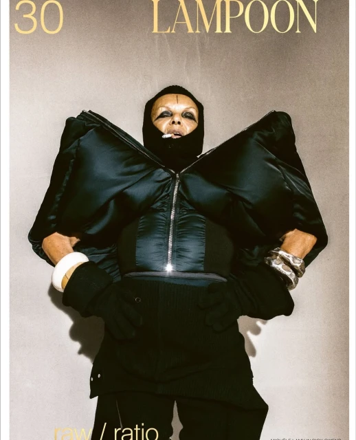 Androgynous person in a black puffer jacket and black pants poses in front of a grey backdrop for a fashion editorial magazine cover.