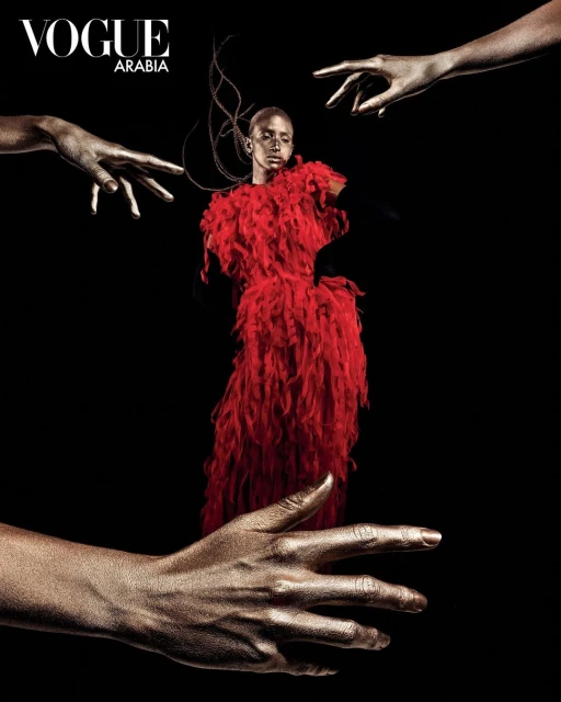 Fashion editorial shot of a bald Black female model in a red feathery dress with gold-painted hands reaching in on a black background.