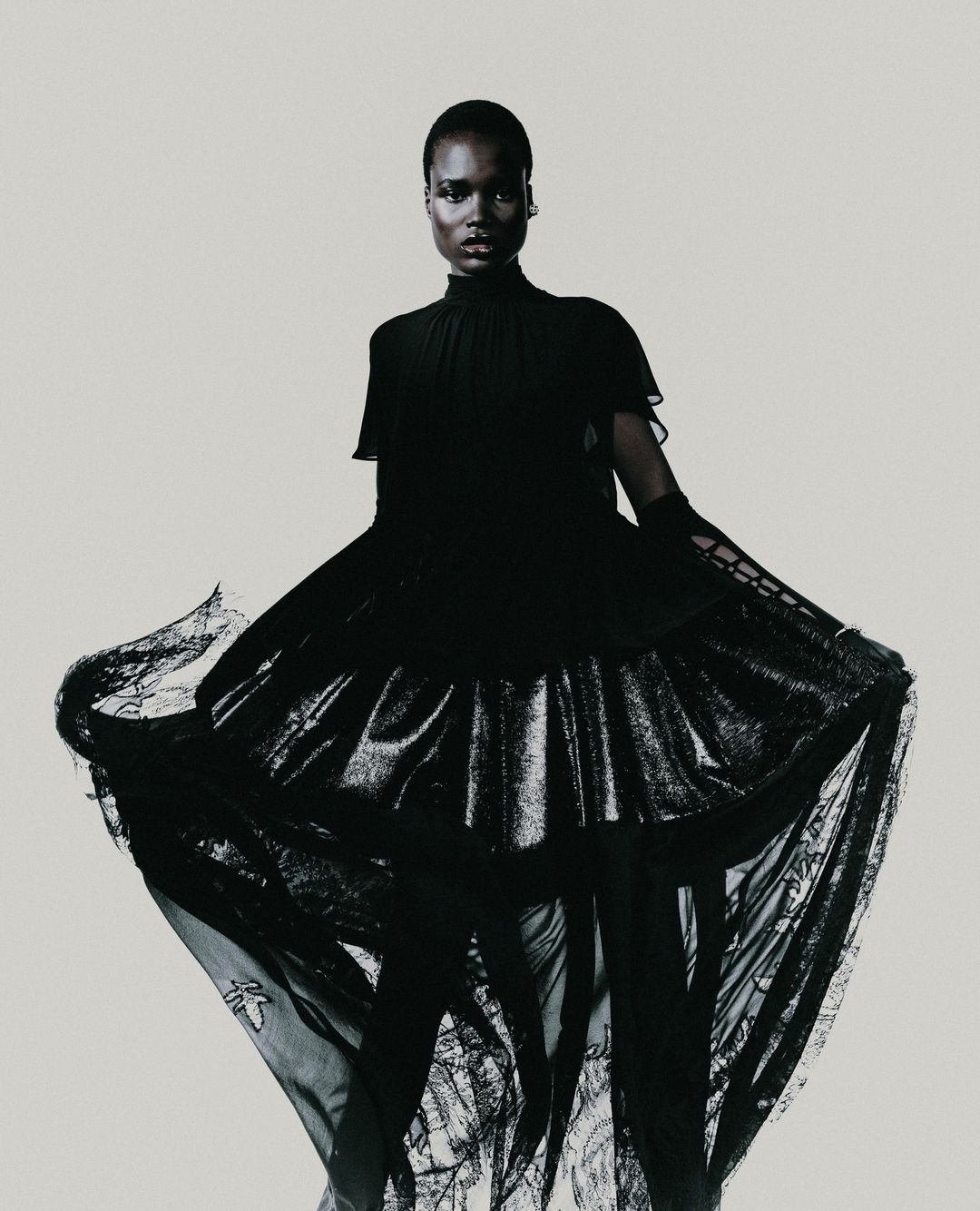 Fashionable woman in long black gown with translucent skirt, posing against a pale backdrop in a studio environment with neutral lighting.