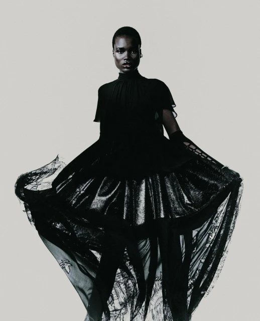 Fashionable woman in long black gown with translucent skirt, posing against a pale backdrop in a studio environment with neutral lighting.