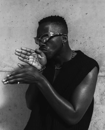 Black man wearing sunglasses and a black vest posing against a concrete wall in a monochrome portrait by Tarek Mawad