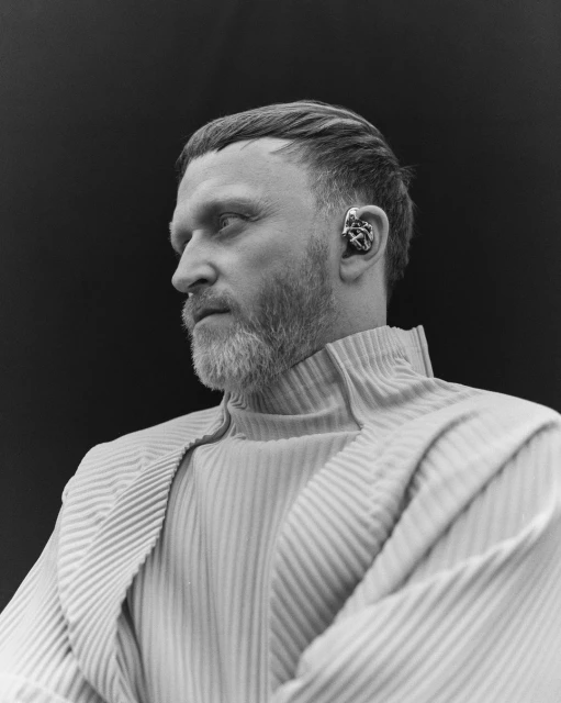 Monochrome portrait of a bearded man with an elaborate ear cuff, wearing a textured, high-necked garment against a dark backdrop.