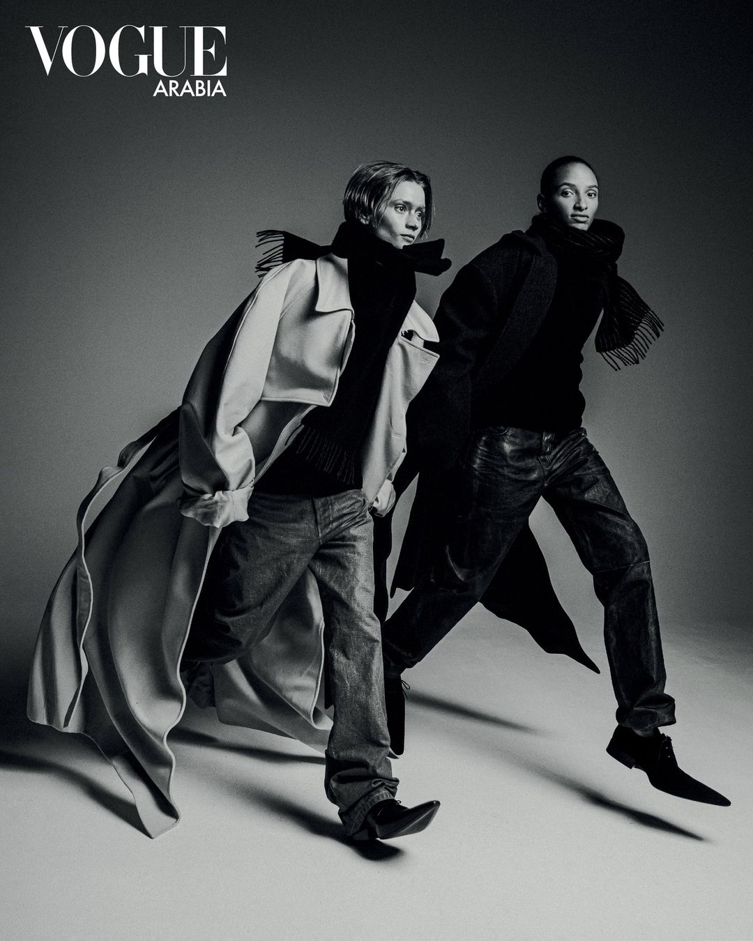Monochrome fashion portrait of two models striding forward against a plain grey studio background.