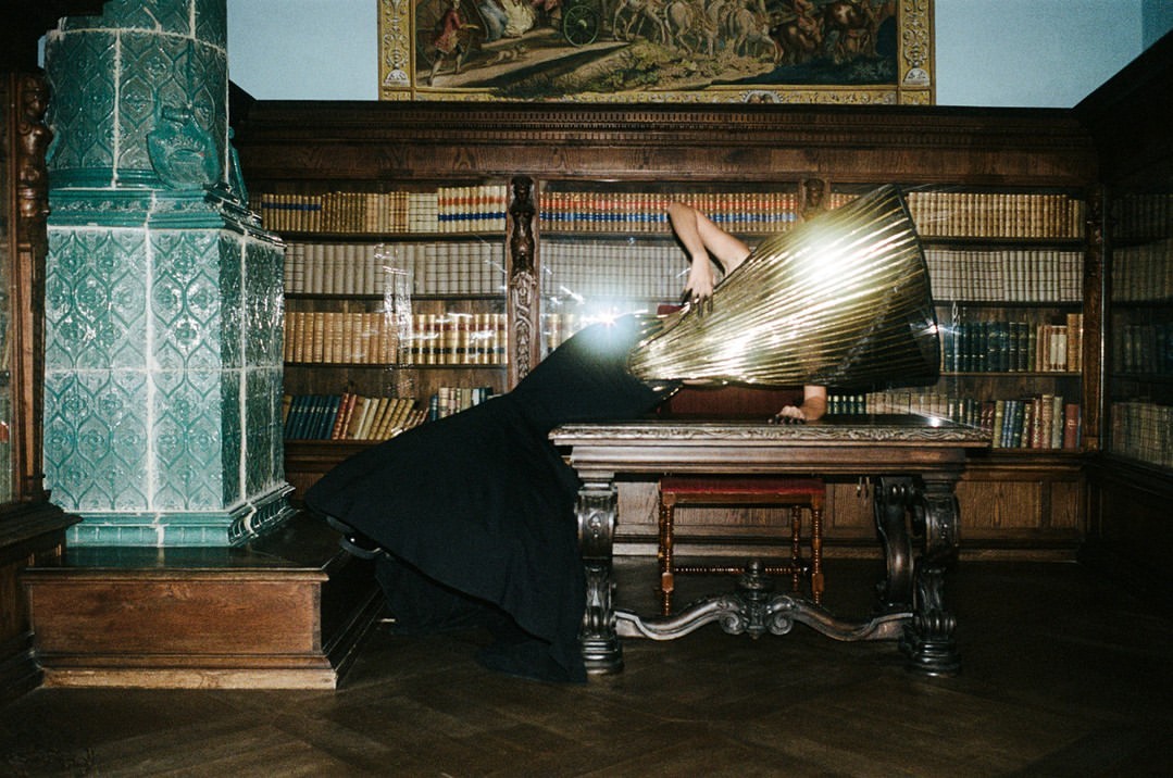 A person in a gold and black dress lying on a table in a library with a tiled fireplace, surrounded by bookshelves and a tapestry.