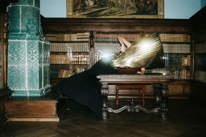 A person in a gold and black dress lying on a table in a library with a tiled fireplace, surrounded by bookshelves and a tapestry.