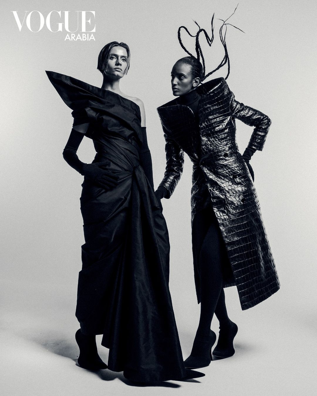 Two fashion models in black haute couture, Vogue Arabia editorial, posing in studio with dramatic shapes and textures in monochrome.