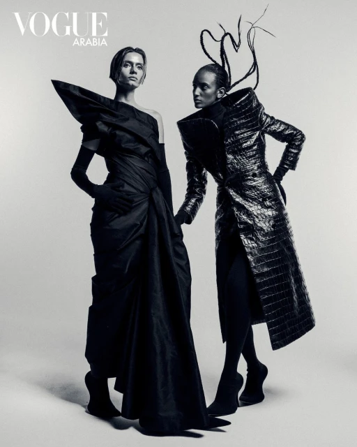 Two fashion models in black haute couture, Vogue Arabia editorial, posing in studio with dramatic shapes and textures in monochrome.