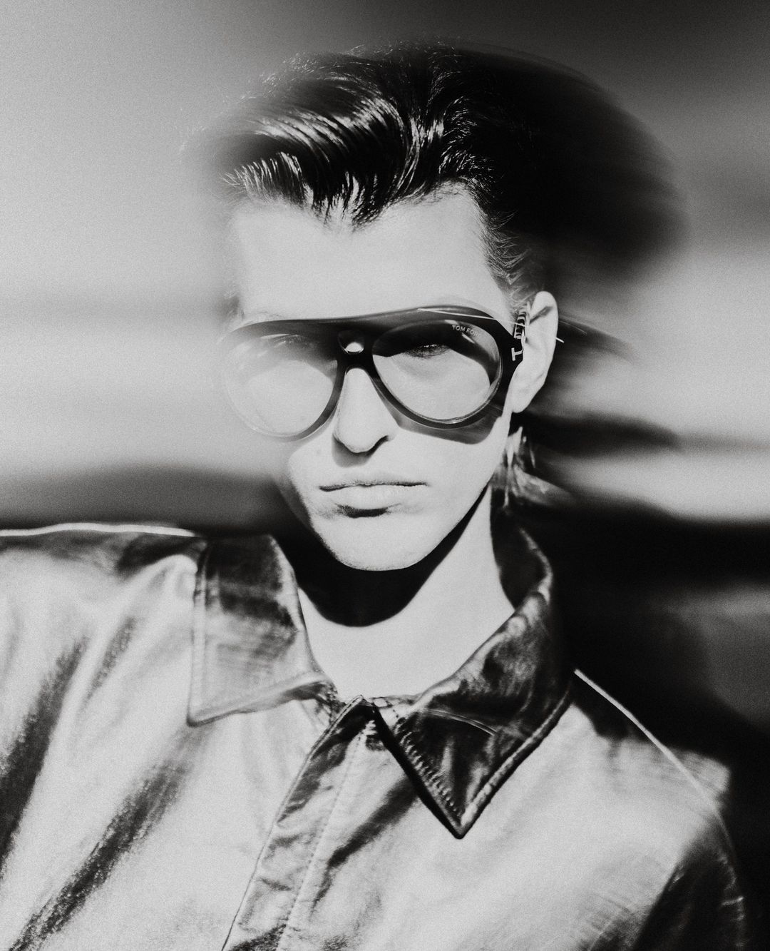 Monochrome portrait of an adult male wearing aviator glasses and a shiny collared shirt against a blurred light background.