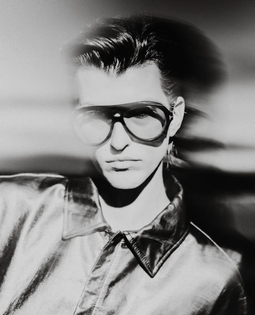 Monochrome portrait of an adult male wearing aviator glasses and a shiny collared shirt against a blurred light background.