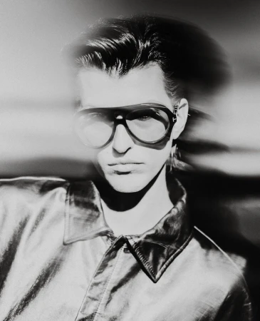 Monochrome portrait of an adult male wearing aviator glasses and a shiny collared shirt against a blurred light background.